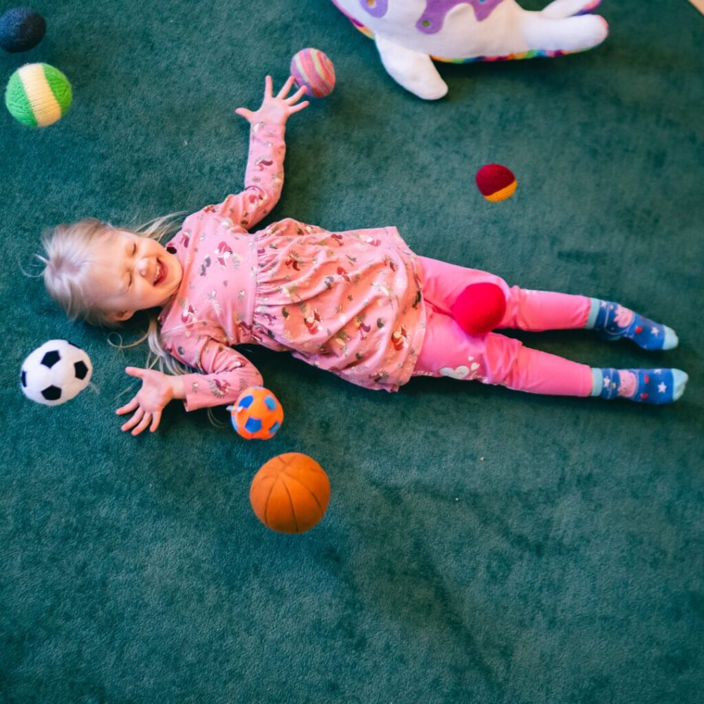 Lapsi makaa vihreällä matolla ympärillään leluja ja palloja päiväkodin leikkihuoneessa.